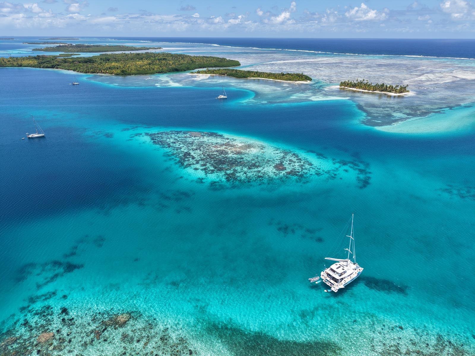 Nicara at anchor over a coral reef beside palm-fringed atoll islands